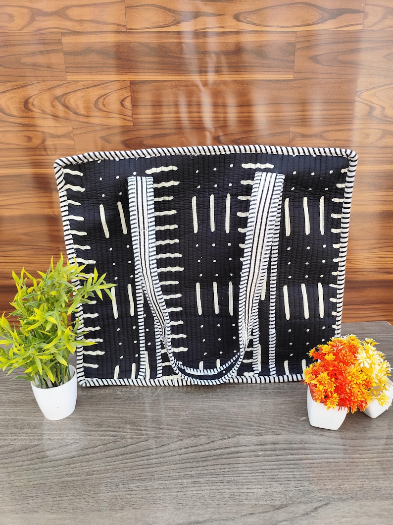 May include: Black tote bag with white vertical and dotted patterns. The bag has white striped handles and trim. The bag is sitting on a wooden surface with small potted plants on either side.