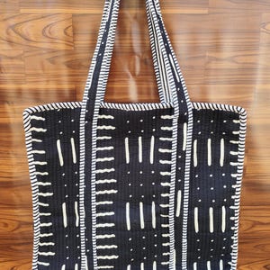 May include: Black and white tote bag with a geometric pattern. The bag features a quilted design with vertical and horizontal white lines and dots on a black background. The handles and edges are trimmed with white and black striped fabric.