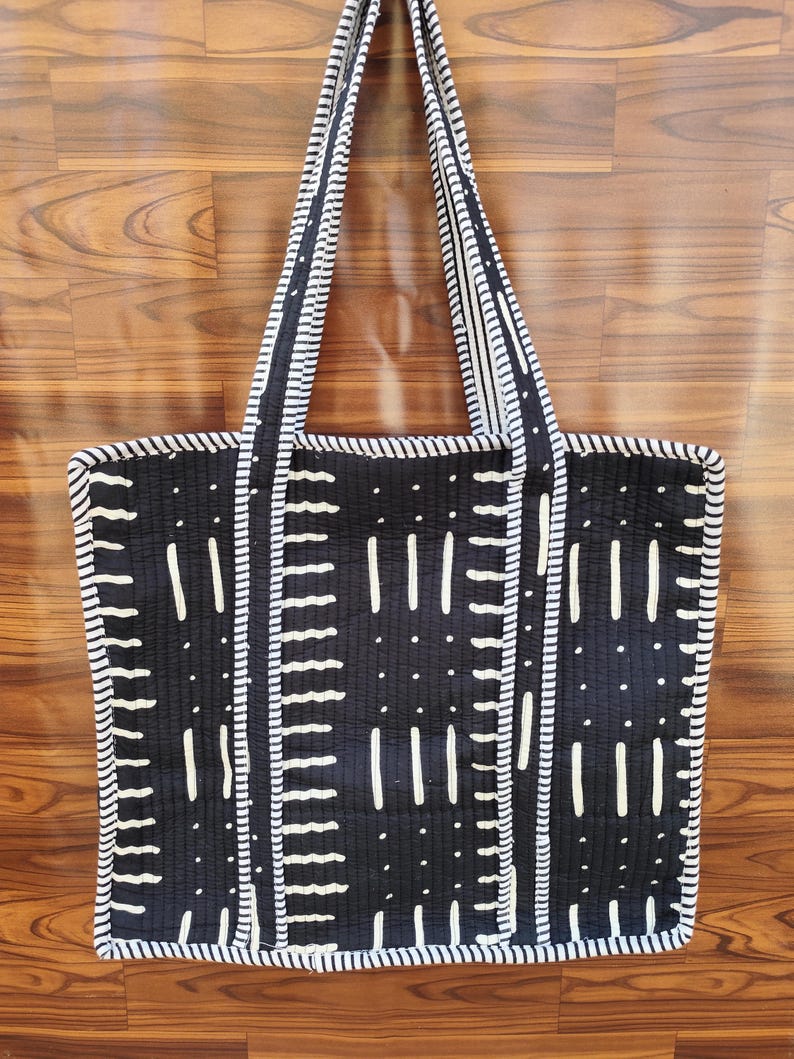 May include: Black and white tote bag with a geometric pattern. The bag features a quilted design with vertical and horizontal white lines and dots on a black background. The handles and edges are trimmed with white and black striped fabric.