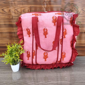May include: Pink tote bag with a lobster print. The bag has burgundy handles and trim, and a ruffled edge. The bag is sitting on a wooden surface. A small potted plant is to the left of the bag.