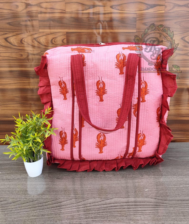May include: Pink tote bag with a lobster print. The bag has burgundy handles and trim, and a ruffled edge. The bag is sitting on a wooden surface. A small potted plant is to the left of the bag.
