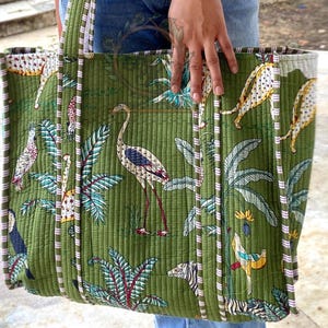 May include: Green quilted tote bag with a tropical print featuring birds, zebras, and leopards. The bag has a striped trim and handles. The bag is held by a person wearing blue jeans.