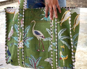 May include: Green quilted tote bag with a tropical print featuring birds, zebras, and leopards. The bag has a striped trim and handles. The bag is held by a person wearing blue jeans.