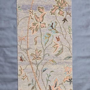 Puede incluir: Alfombra rectangular con estampado floral sobre fondo lila claro. El diseño presenta flores y hojas estilizadas en tonos rosa, azul y verde, con tallos marrones. La alfombra tiene un borde con flecos.