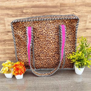 Könnte beinhalten: Eine rechteckige Tragetasche mit Leopardenmuster. Die Tasche hat rosa Griffe und ist mit schwarz-weißen Streifen verziert. Die Tasche steht auf einer Holzoberfläche, mit kleinen Topfpflanzen davor.