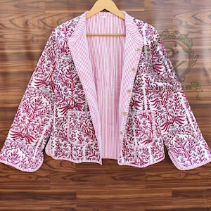 Könnte beinhalten: Eine gesteppte Jacke mit floralem Muster in Rosa- und Weißtönen. Die Jacke hat ein gestreiftes rosa Futter und zwei Vordertaschen. Sie hängt an einem Holzbügel.