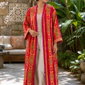 Op de afbeelding: Een lange, aan de voorzijde open kimono met een levendig patroon van oranje en gele tijgermotieven op een felroze achtergrond. Het kledingstuk heeft wijde mouwen met gestreepte boorden en een effen roze bies langs de opening.
