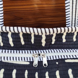 May include: Close-up of a black and white patterned tote bag. The bag features a white zipper and two silver zipper pulls. The bag has a quilted design with white vertical dashes on a black background.