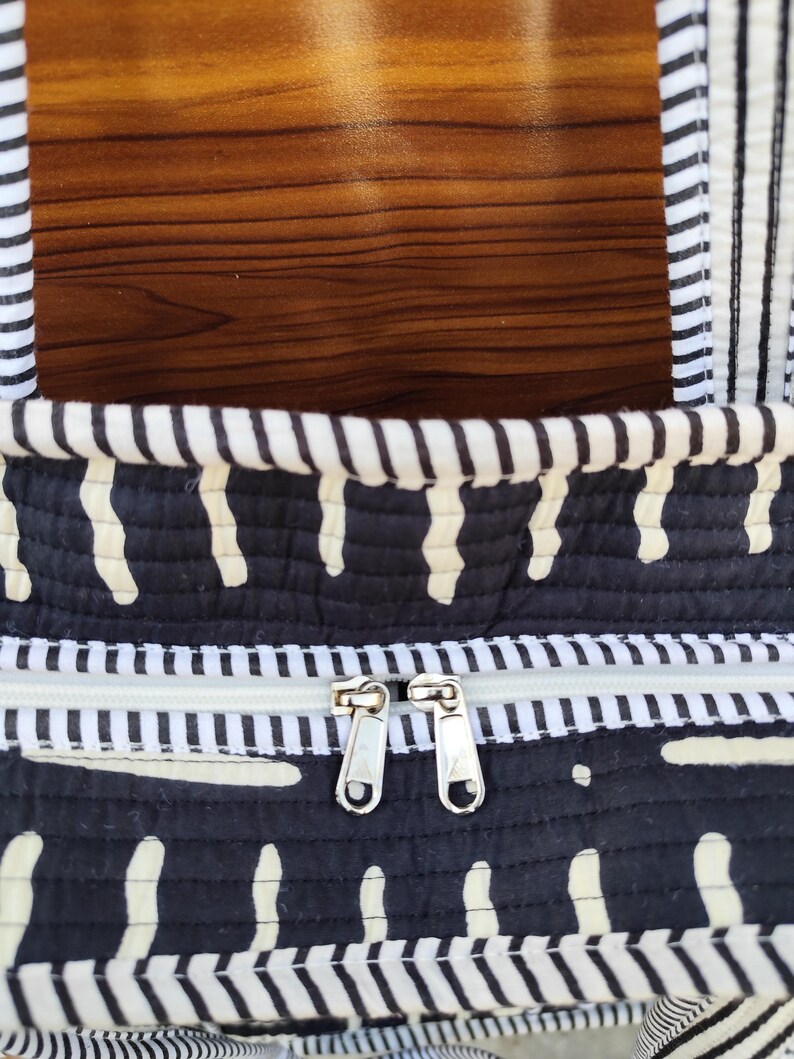 May include: Close-up of a black and white patterned tote bag. The bag features a white zipper and two silver zipper pulls. The bag has a quilted design with white vertical dashes on a black background.