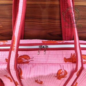 May include: Pink quilted tote bag with a repeating orange lobster pattern. The bag has red trim, handles, and a zipper closure. The background is a wooden surface.