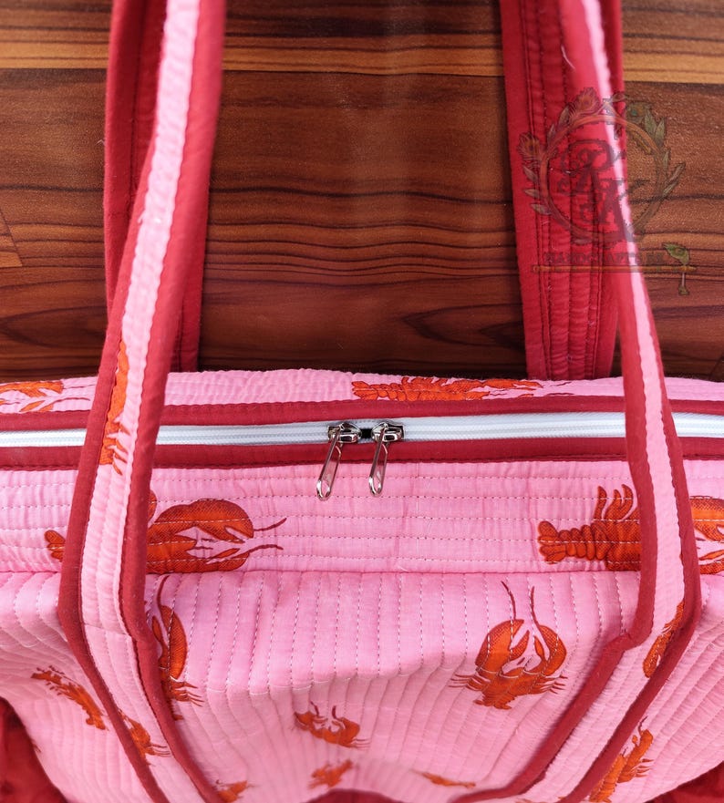 May include: Pink quilted tote bag with a repeating orange lobster pattern. The bag has red trim, handles, and a zipper closure. The background is a wooden surface.
