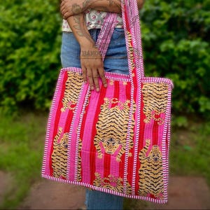 May include: A quilted tote bag with a tiger print. The bag features a repeating pattern of tigers in gold and black stripes against a pink and red striped background. The bag has a rectangular shape with matching handles.