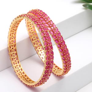 May include: Two gold bangles adorned with pink gemstones. The gemstones are oval-shaped and closely set, forming a continuous pattern. The bangles are presented on a white surface, highlighting their intricate design. The overall aesthetic is one of elegance.
