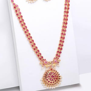 Puede incluir: Un conjunto de joyas dorado con un collar y pendientes a juego. El collar tiene un diseño largo de doble hebra con piedras ovaladas de color rubí. El colgante y los pendientes tienen un diseño floral con piedras de color rubí y pequeñas perlas blancas.
