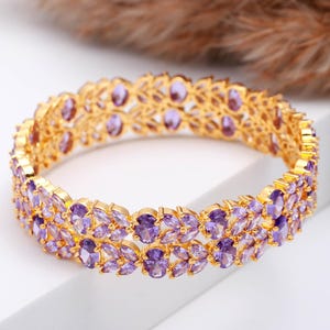 Puede incluir: Un brazalete dorado con dos filas de piedras preciosas ovaladas de color púrpura. El brazalete tiene un diseño calado intrincado. Las piedras preciosas son de un color púrpura vibrante, sobre un fondo blanco.