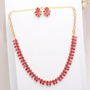 Puede incluir: Un conjunto de collar y pendientes a juego en tono dorado. El collar y los pendientes están adornados con vibrantes piedras preciosas rojas. El collar presenta una delicada cadena con un patrón repetido de piedras preciosas rojas. Los pendientes tienen un racimo de las mismas piedras preciosas rojas.