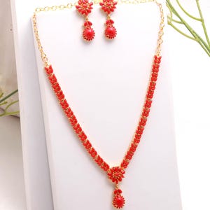 Puede incluir: Un conjunto de joyas rojo y dorado con un collar y pendientes a juego. El collar tiene un colgante floral y un diseño en forma de hoja. Los pendientes tienen una parte superior floral y una piedra roja en forma de lágrima. El conjunto se exhibe sobre una superficie blanca.