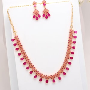 May include: A gold-toned necklace and earring set. The necklace and earrings feature a series of small, pink gemstones and larger, teardrop-shaped pink gemstones. The jewelry is displayed on a white surface.