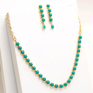 Op de afbeelding: Een goudkleurige ketting en bijpassende oorbellen set. De ketting en oorbellen zijn voorzien van turquoise, ronde edelstenen. De oorbellen hebben een druppelvormige edelsteen aan de onderkant. De sieraden worden gepresenteerd op een wit oppervlak.