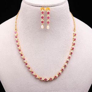 May include: A gold necklace and earring set. The necklace and earrings feature alternating red gemstones and small white accents. The earrings have a teardrop-shaped pearl at the bottom. The jewellery is displayed on a mannequin.
