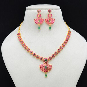 Peut inclure: Un ensemble de bijoux dorés comprenant un collier et des boucles d'oreilles assorties. Le collier et les boucles d'oreilles sont ornés de pierres précieuses roses et vertes, disposées en motif floral. L'ensemble est présenté sur un mannequin blanc sur fond noir.