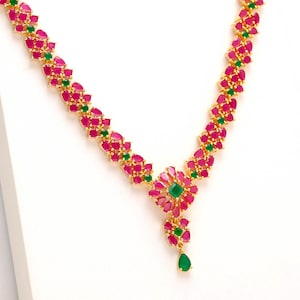 Pode incluir: Um colar dourado com um design floral com pedras preciosas vermelho rubi e verde esmeralda. O colar tem um pingente central com um aglomerado em forma de flor de pedras vermelhas e uma pedra central verde quadrada.