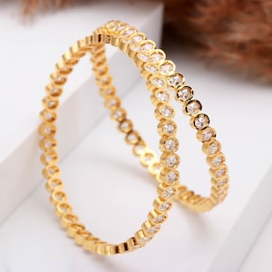 Peut inclure: Deux bracelets dorés ornés d'un motif répété de pierres précieuses rondes et transparentes. Les bracelets sont présentés sur une surface blanche, avec un arrière-plan flou brun.