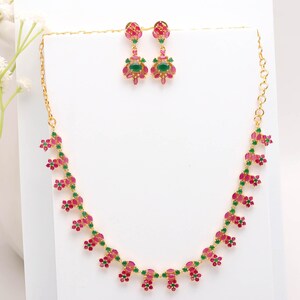 Puede incluir: Un conjunto de collar y pendientes dorados. La joyería presenta un diseño floral con piedras preciosas rojas y verdes. Los pendientes son de tipo botón. El collar y los pendientes se exhiben sobre una superficie blanca.