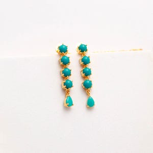 Op de afbeelding: Een paar goudkleurige oorhangers. Elke oorbel heeft een reeks turquoise kralen, die uitmonden in een druppelvormige turquoise kraal. De oorbellen staan tegen een witte achtergrond.