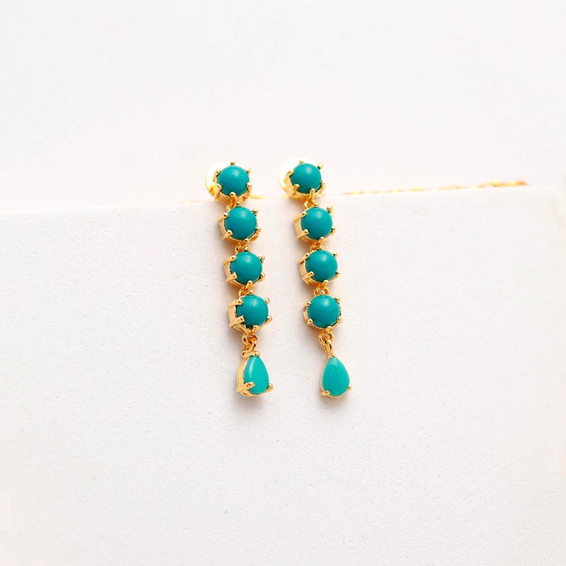Op de afbeelding: Een paar goudkleurige oorhangers. Elke oorbel heeft een reeks turquoise kralen, die uitmonden in een druppelvormige turquoise kraal. De oorbellen staan tegen een witte achtergrond.