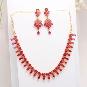 May include: A gold-toned jewelry set featuring a necklace and matching earrings. The necklace and earrings are adorned with numerous small, deep pink gemstones. The earrings have a chandelier design, and the necklace has a curved shape.