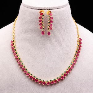Puede incluir: Un conjunto de collar y pendientes dorados. La joyería presenta un patrón repetido de piedras rojas en forma de lágrima, intercaladas con pequeñas piedras verdes. Los pendientes cuelgan y el collar está sobre un maniquí.