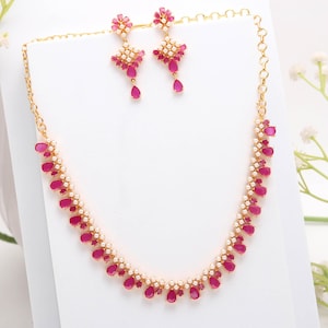 Puede incluir: Un conjunto de joyas dorado que incluye un collar y pendientes a juego. El collar y los pendientes están adornados con piedras preciosas rojas en forma de lágrima y pequeñas perlas blancas. Los pendientes tienen un diseño floral en la parte superior. El conjunto se exhibe sobre una superficie blanca.