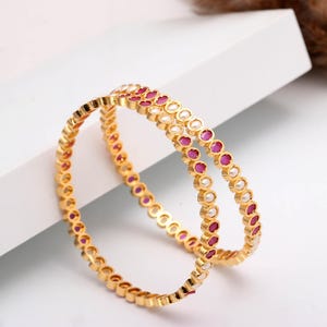 Könnte beinhalten: Zwei goldene Armreifen mit abwechselnden Rubinen und perlenartigen Akzenten. Die Armreifen werden auf einer weißen Oberfläche präsentiert, um ihr detailliertes Design und ihre leuchtenden Farben hervorzuheben. Ein zeitloses Schmuckstück.