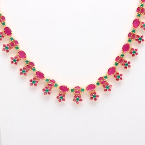 Op de afbeelding: Een goudkleurige ketting met ovale, robijnkleurige edelstenen en kleinere, bloemvormige clusters van groene en rode stenen. De ketting is geplaatst tegen een witte achtergrond, die de levendige kleuren en het ingewikkelde ontwerp benadrukt.