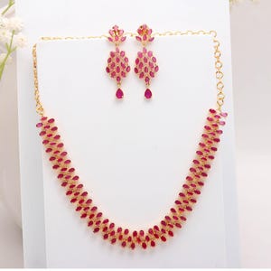 Puede incluir: Un conjunto de collar y pendientes dorados. La joyería presenta un patrón de pequeñas piedras ovaladas de color rubí. Los pendientes son de estilo colgante, con una sola lágrima de color rubí en la parte inferior. El collar mide unos 40 cm.