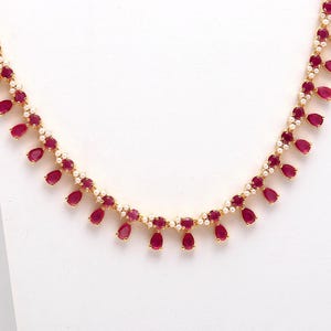 Może przedstawiać: Złoty naszyjnik z naprzemiennymi czerwonymi kamieniami w kształcie łezki i małymi białymi perłami. Naszyjnik jest prezentowany na białym tle, podkreślając wzór i kolor biżuterii.