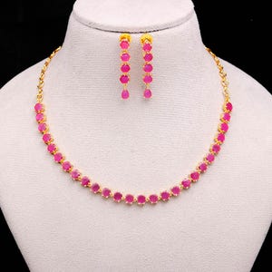 May include: A gold-coloured necklace and earring set. The necklace and earrings feature a series of round, bright pink gemstones. The earrings are dangling, and the necklace sits on a white mannequin. The jewellery is displayed against a black background.