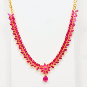 Puede incluir: Un collar dorado con un colgante floral y un dije en forma de lágrima. El collar está adornado con gemas rosas vibrantes dispuestas en un patrón de hojas. El collar se exhibe sobre un fondo blanco.