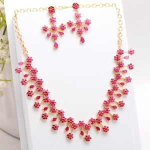 Puede incluir: Un conjunto de collar y pendientes de color dorado. El collar y los pendientes presentan un diseño floral con pequeñas piedras preciosas rosas. Los pendientes tienen forma de cruz con gemas colgantes en forma de lágrima. El collar tiene un diseño similar.