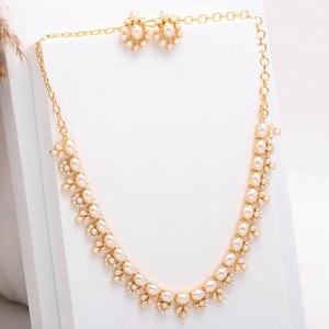 May include: A gold-coloured necklace and matching earrings set. The necklace and earrings feature off-white pearls and small, clear crystals. The jewellery is displayed on a white surface, highlighting the elegant design.