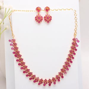 May include: A gold-coloured necklace and matching earrings set. The necklace and earrings feature a floral design with vibrant pink gemstones. The earrings are stud style, and the necklace drapes gracefully. The jewellery is displayed on a white surface.