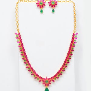 May include: A gold-colored necklace and earring set. The necklace and earrings feature a floral design with pink and green gemstones. The necklace has a teardrop-shaped green gemstone pendant. The earrings have a matching teardrop-shaped green gemstone.