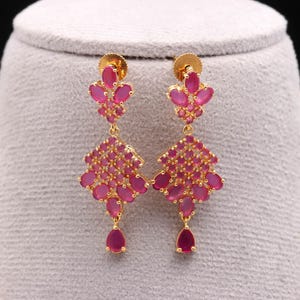 May include: Gold-toned earrings with a floral design, featuring clusters of pink gemstones. The earrings have a stud at the top, a floral cluster in the middle, and a teardrop-shaped gemstone at the bottom. The gemstones are a vibrant pink color.