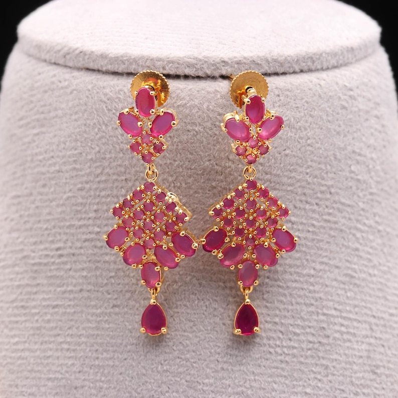 May include: Gold-toned earrings with a floral design, featuring clusters of pink gemstones. The earrings have a stud at the top, a floral cluster in the middle, and a teardrop-shaped gemstone at the bottom. The gemstones are a vibrant pink color.