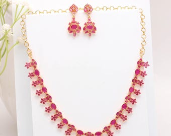 May include: A gold-toned necklace and earring set featuring ruby-colored gemstones. The necklace and earrings have a floral design with oval and flower-shaped stones. The jewelry is displayed on a white surface.