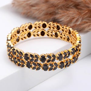 Könnte beinhalten: Ein goldfarbenes Armband mit zwei Reihen schwarzer Edelsteine. Das Armband hat ein aufwendiges Design mit oval geformten schwarzen Steinen, die in einen goldfarbenen Metallrahmen gefasst sind. Der Schmuck wird auf einer weißen Oberfläche präsentiert.
