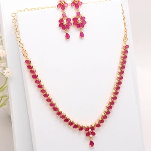 Puede incluir: Un conjunto de collar y pendientes a juego en tono dorado. La joyería presenta piedras en forma de lágrima de color rubí y pequeños detalles blancos. Los pendientes tienen un diseño floral con gotas colgantes. El collar tiene un diseño similar.