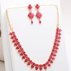 May include: A gold-toned jewelry set featuring a necklace and matching earrings. The necklace and earrings are adorned with vibrant pink gemstones arranged in a floral pattern. The earrings have a teardrop-shaped gemstone at the bottom.