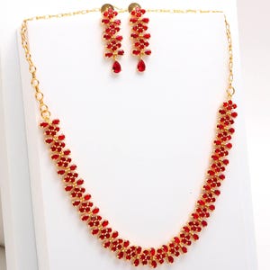 Puede incluir: Un conjunto de joyas de color dorado que incluye un collar y pendientes a juego. El collar y los pendientes están adornados con piedras preciosas rojas vibrantes dispuestas en un patrón floral. Los pendientes tienen una gema en forma de lágrima.